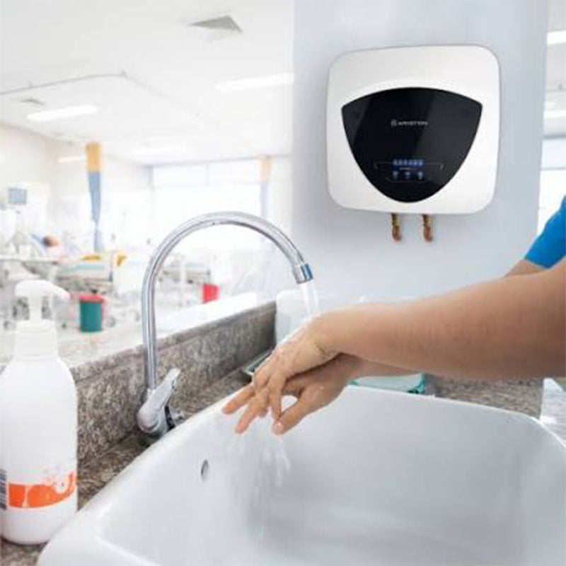A hand-washing station in a bright bathroom or clinic setting, showing a person's hands being washed under a running faucet with a wall-mounted water heater above.
