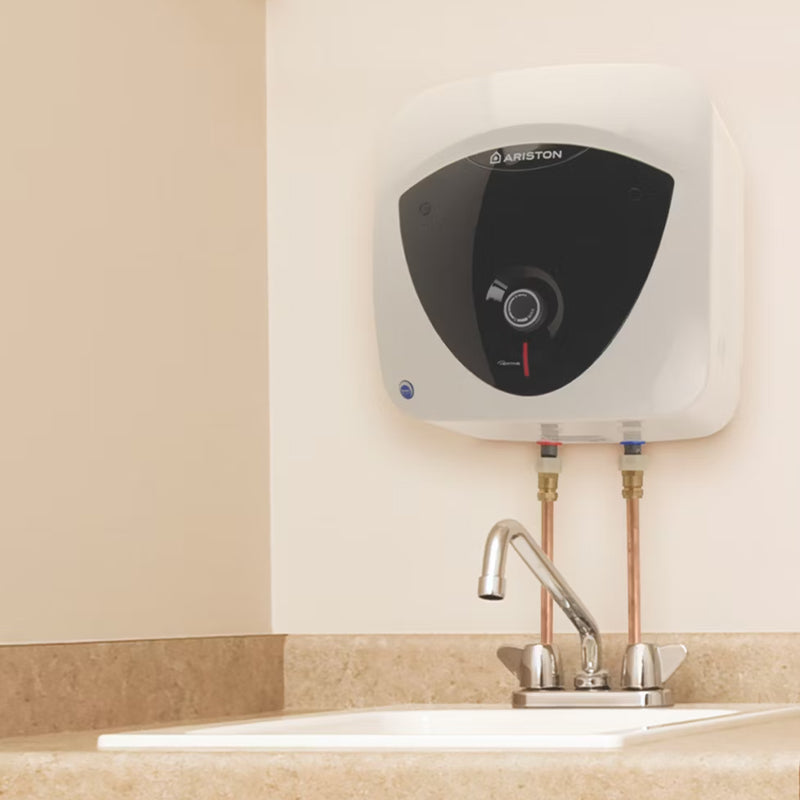 Ariston wall-mounted water heater in white with a black front panel, installed above a sink with a chrome faucet on a beige tiled wall.
